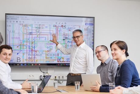 Ein Team diskutiert im Besprechungsraum vor einem großen Bildschirm einen technischen Plan.
