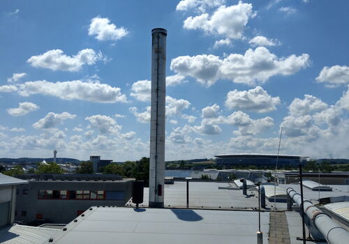 Ein hoher Schornstein ragt über die Industriegebäude, während am Himmel weiße Wolken vorüberziehen.