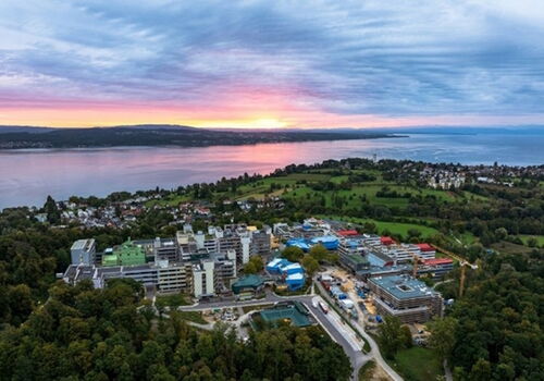 Luftaufnahme eines Campus am Bodensee bei Sonnenuntergang, umgeben von grünen Wiesen und bewaldeten Hügeln.