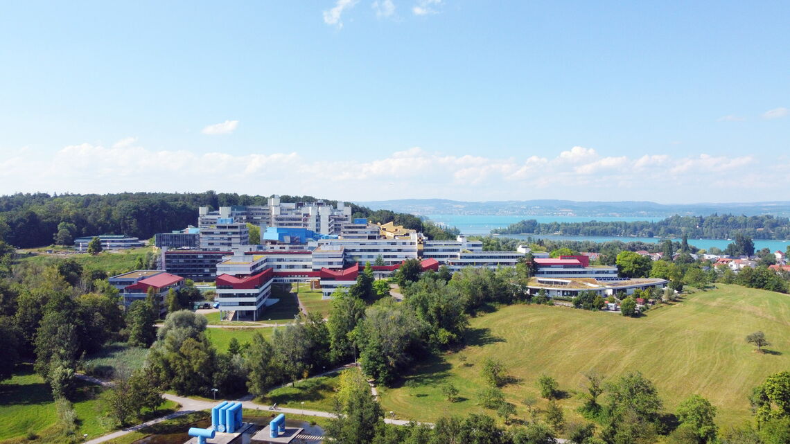 Ein modernes Universitätsgebäude liegt inmitten grüner Landschaft mit Blick auf einen malerischen See.