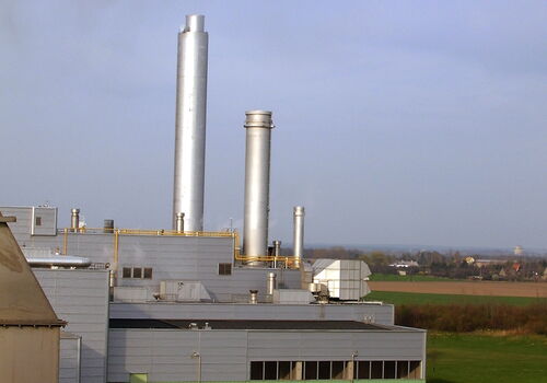 Eine moderne Fabrik mit hohen Schornsteinen steht inmitten einer grünen Landschaft unter bewölktem Himmel.