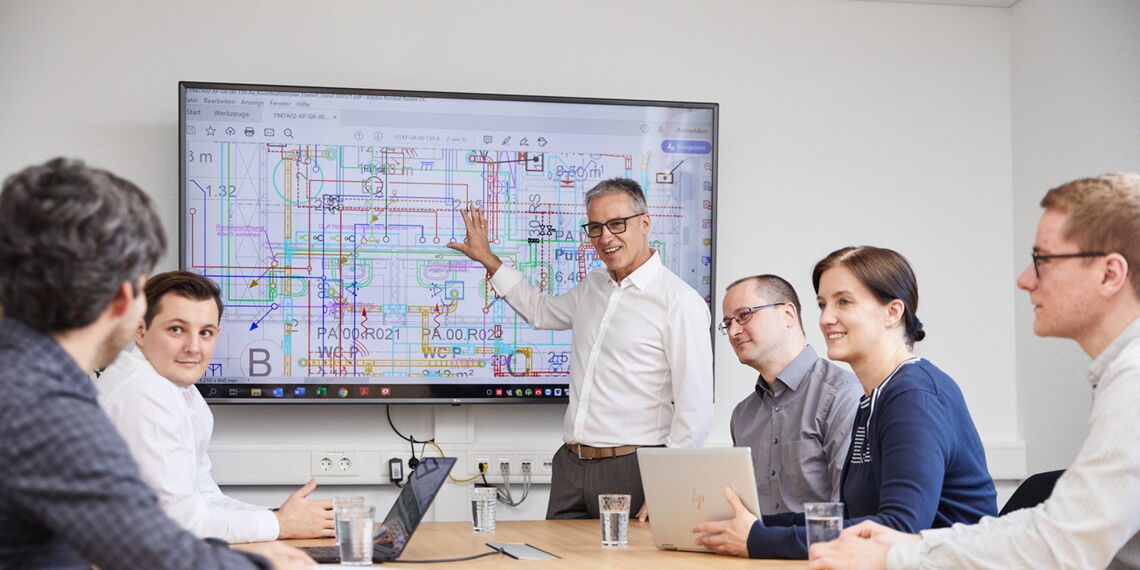 Ein Team diskutiert im Besprechungsraum vor einem großen Bildschirm einen technischen Plan.