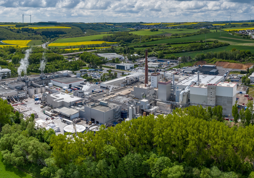 Eine große Industrieanlage raucht inmitten grüner Landschaft, umgeben von Feldern und Hügeln.